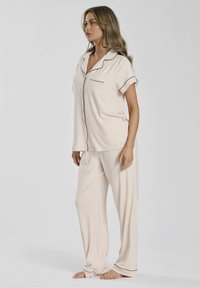 Beige pyjama set van zachte stof, met een kort mouwtop met kraag en zwarte piping, en een zak, gecombineerd met losse broek.