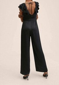 Femme portant une combinaison noire à jambes larges avec des manches volantées et un dos plongeant en V noué avec de fines bretelles, associée à des sandales à talons noires.