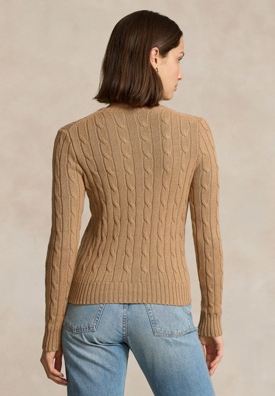 Sieviete ar īsiem brūniem matiem, ģērbusies pieguļošā kamieļa krāsas adītā džemperī ar pinumu rakstu un gaiši zilās džinsās, skatās prom pret neitrālu fonu.