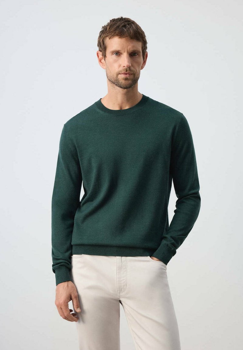 Homme aux cheveux courts et bruns portant un pull vert foncé à manches longues et un pantalon beige clair, debout devant un fond clair uni.
