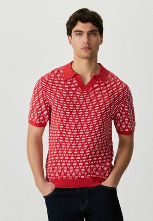 Jeune homme aux cheveux foncés portant un polo à manches courtes rouge et blanc à motifs, debout les mains dans les poches de son pantalon foncé.