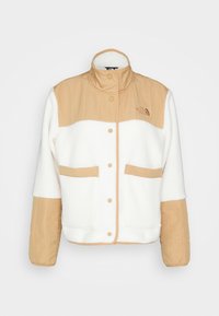 Fleece-takki, jossa on vaaleanbeigejä ja valkoisia väri-iloituksia, korkea kaulus, painonapit ja kaksi etutaskua. Kirjailtu logoyksityiskohta.