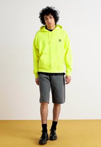 Neon yellow hooded sweatshirt with a front pocket, paired with gray shorts. Black socks and shoes complete the outfit. Simple design, no patterns.