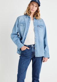 Lyseblå denimskjorte med trykknapper og to brystlommer, kombinert med mørkeblå jeans som har en tettsittende design og subtil tekstur.