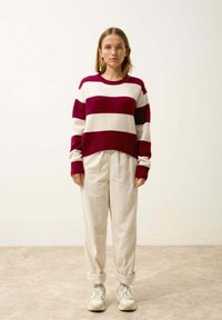 Pull rayé bordeaux et crème, associé à un pantalon blanc texturé et des baskets claires. Le pull possède un col rond et des poignets côtelés.