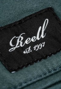 Etiqueta de tela negra con texto bordado en blanco "Reell" y "est. 1997," cosida sobre un material texturizado en verde azulado con detalles de costura visibles.