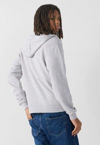 Sudadera de algodón gris con capucha ajustable, corte relajado y puños acanalados, llevada con jeans azules, mostrando la vista desde atrás.