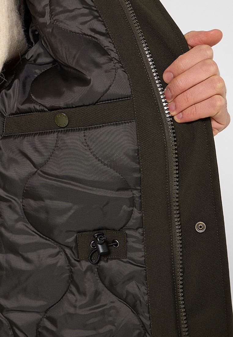 Intérieur de veste vert olive avec doublure matelassée noire, fermeture éclair et détail à bouton-pression. Comprend un cordon de serrage pour l'ajustement.