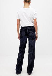 Merenneidon samettihousut leveällä lahkeella, joissa on teksturoitu pinta ja hienovaraisia muunnelmia kangaskuviossa, yhdistettynä valkoiseen paitaan.