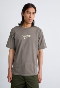 En person står mod en lys baggrund iført en grå t-shirt med et Nike-logo og grønne bukser, mens de kigger direkte ind i kameraet.