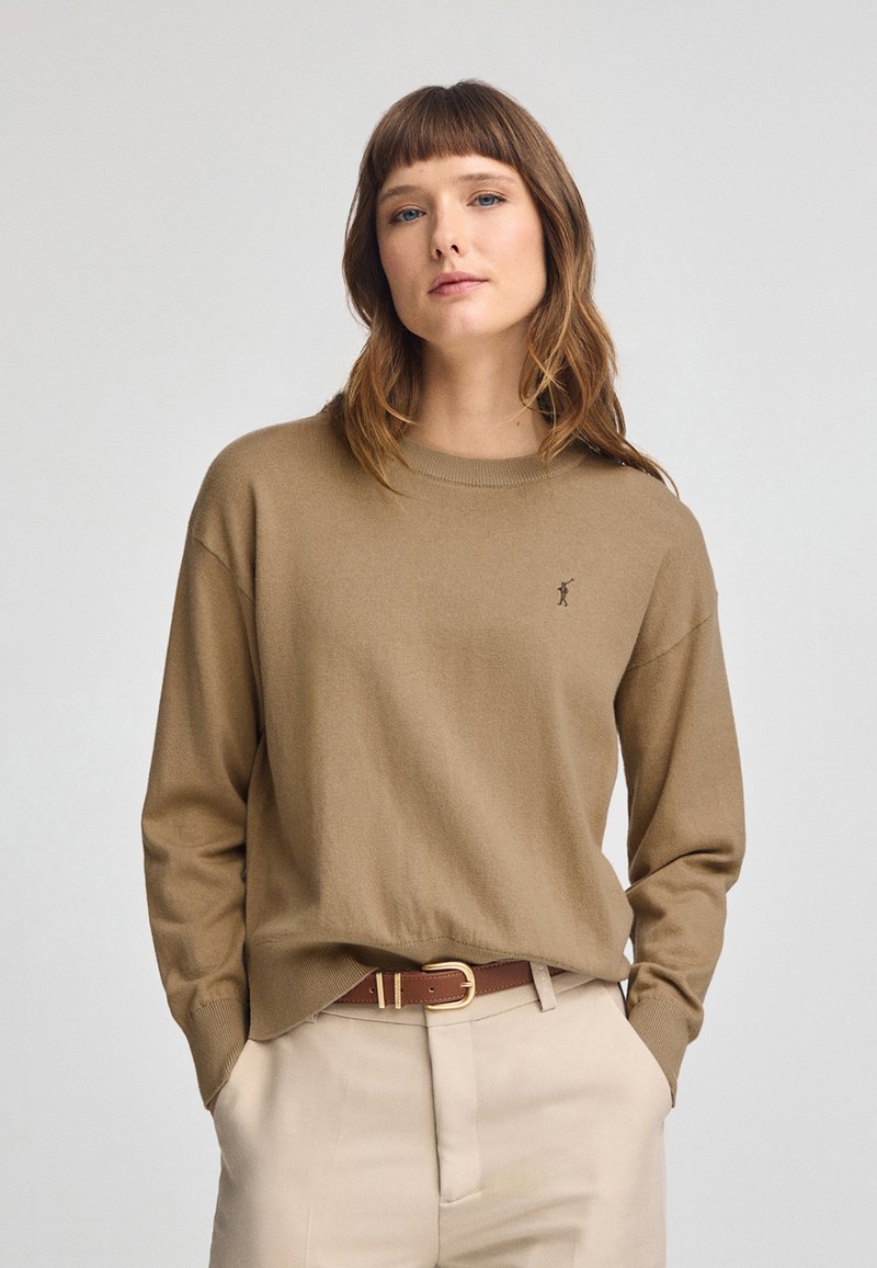Brauner Strickpullover mit Rundhalsausschnitt und langen Ärmeln, mit einem kleinen Logo auf der Brust. Kombiniert mit hellbeigen Hosen und einem Gürtel.
