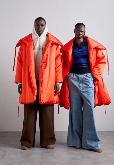 Δύο μοντέλα φοράνε υπερμεγέθη, φωτεινά πορτοκαλί фουσκωτά μπουφάν με μεγάλα καπέλα. Ο άντρας το συνδυάζει με μια γκρι φούτερ και καφέ παντελόνι με φαρδιά γραμμή, ενώ η γυναίκα φοράει ένα μπλε ριγέ πουλόβερ και ανοιχτόχρωμα τζιν.