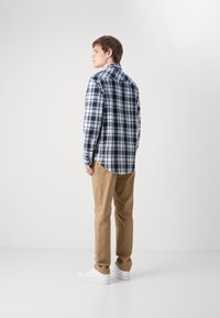 Camisa de cuadros para hombre en navy, blanco y azul claro. Tela de algodón, botón frontal, mangas largas, dobladillo redondeado. Combinada con pantalones beige y zapatos blancos.