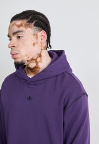Lilla hettegenser med snor og svart Adidas-logo, med en myk tekstur og uformell design, båret av en person med stylet hår.