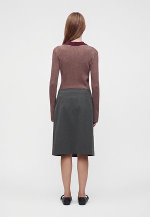 Femme aux cheveux longs et raides bruns portant un haut rose à manches longues, une jupe grise longueur genoux et des chaussures plates noires, debout de dos.