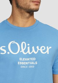 Světle modré bavlněné tričko s kulatým výstřihem. Na přední straně se nachází výrazný bílý text "s.Oliver ELEVATED ESSENTIALS SINCE 1969".