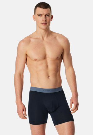 Oberkörperfreier Mann mit kurzen Haaren, der marineblaue Boxershorts mit einem grauen Bund vor einem schlichten hellen Hintergrund trägt.