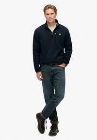 Marineblauer Fleece-Pullover mit Reißverschlusskragen, getragen über einem hellen Hemd, kombiniert mit blauen Jeans und schwarzen Schnürstiefeln. Auffälliges Logo auf der Brust.