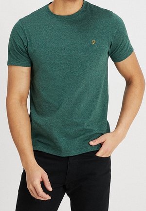 Uomo che indossa una maglietta verde aderente a maniche corte con un piccolo logo giallo sul petto sinistro e pantaloni neri.