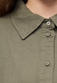 Κοντινό πλάνο ενός πουκαμίσου χακί με γιακά και τρία ορατά κουμπιά, που αναδεικνύει υφή και λεπτομέρειες ραφής.