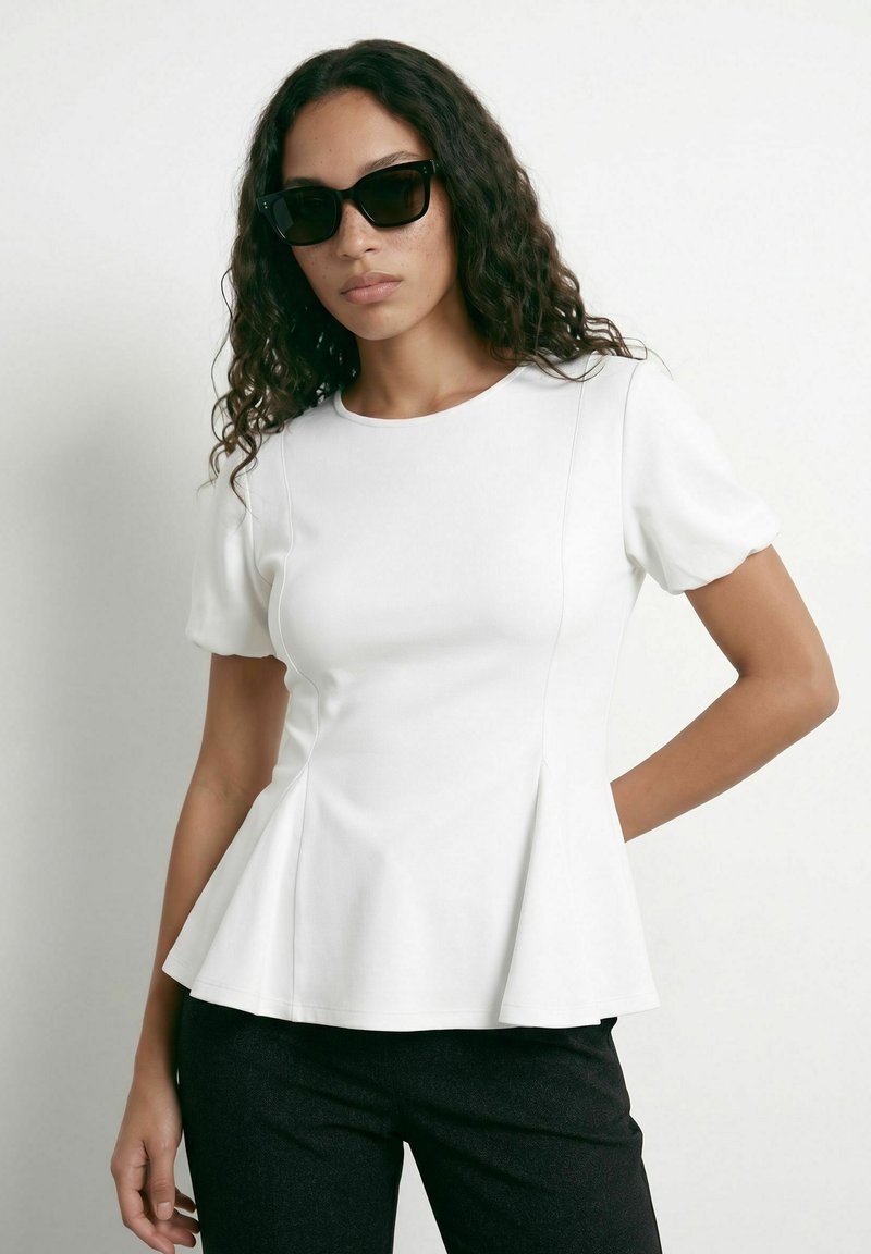 Femme aux cheveux bouclés portant des lunettes de soleil noires, un haut péplum blanc à manches courtes et un pantalon noir, debout devant un fond uni.