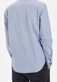 Vaaleansininen nappikauluspaida, jossa on pieni pilkullinen kuvio. Pitkät hihat, joissa on napit, sileä rakenne ja pyöristetty helma.