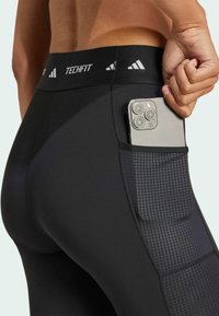 Leggings Techfit negros con un bolsillo lateral texturizado, sosteniendo un smartphone. La tela es suave y elástica con una cinturilla ancha para ofrecer soporte.