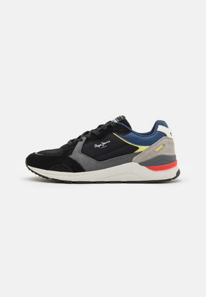 Zapatilla deportiva negra y gris con materiales de ante y malla, acentos contrastantes en azul y amarillo, y una suela intermedia blanca con detalles rojos.