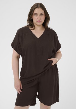 Młoda kobieta o włosach do ramion, ubrana w luźną ciemnobrązową bluzkę z dekoltem w serek i pasujące szorty, stoi z jedną ręką w kieszeni.
