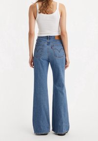 Jean évasé bleu en denim avec un lavage clair. Il présente une étiquette en cuir marron à l'arrière et un design de couture distinctif sur les poches arrière.