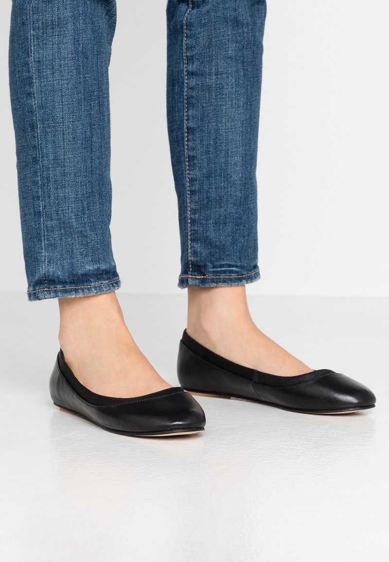 Ballerines en cuir noir avec un bout arrondi et un bord élastique, associées à un jean bleu retroussé, sur fond blanc.