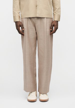 Άνδρας που φοράει μπεζ παντελόνι φαρδιάς γραμμής, κρεμ αθλητικά παπούτσια και μπεζ πουκάμισο με κουμπιά, στέκεται μπροστά σε απλό λευκό φόντο.