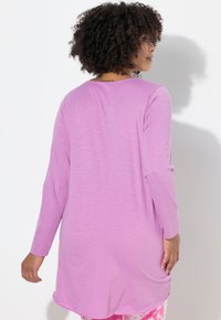 Pitkähihainen, vaaleanvioletti tunika pyöreällä helmalla. Pehmeä, teksturoitu kangas. Mallin selkä näkyvissä, hänellä on kontrastiset kuvioidut legginsit.