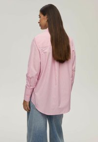 Jasnoróżowa koszula w paski o luźnym kroju z długimi rękawami i zaokrąglonym dołem; połączona z luźnymi niebieskimi dżinsami. Materiał bawełniany, gładka tekstura.