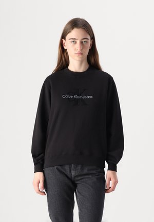 Sweatshirt noir en tissu doux avec des poignets et un ourlet côtelés, arborant un logo "Calvin Klein Jeans" brodé en blanc. Coupe décontractée.