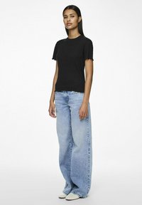 Svart t-skjorte med korte ermer og frynsete kanter, parret med lyseblå vide jeans. Modellen står foran en enkel hvit bakgrunn.