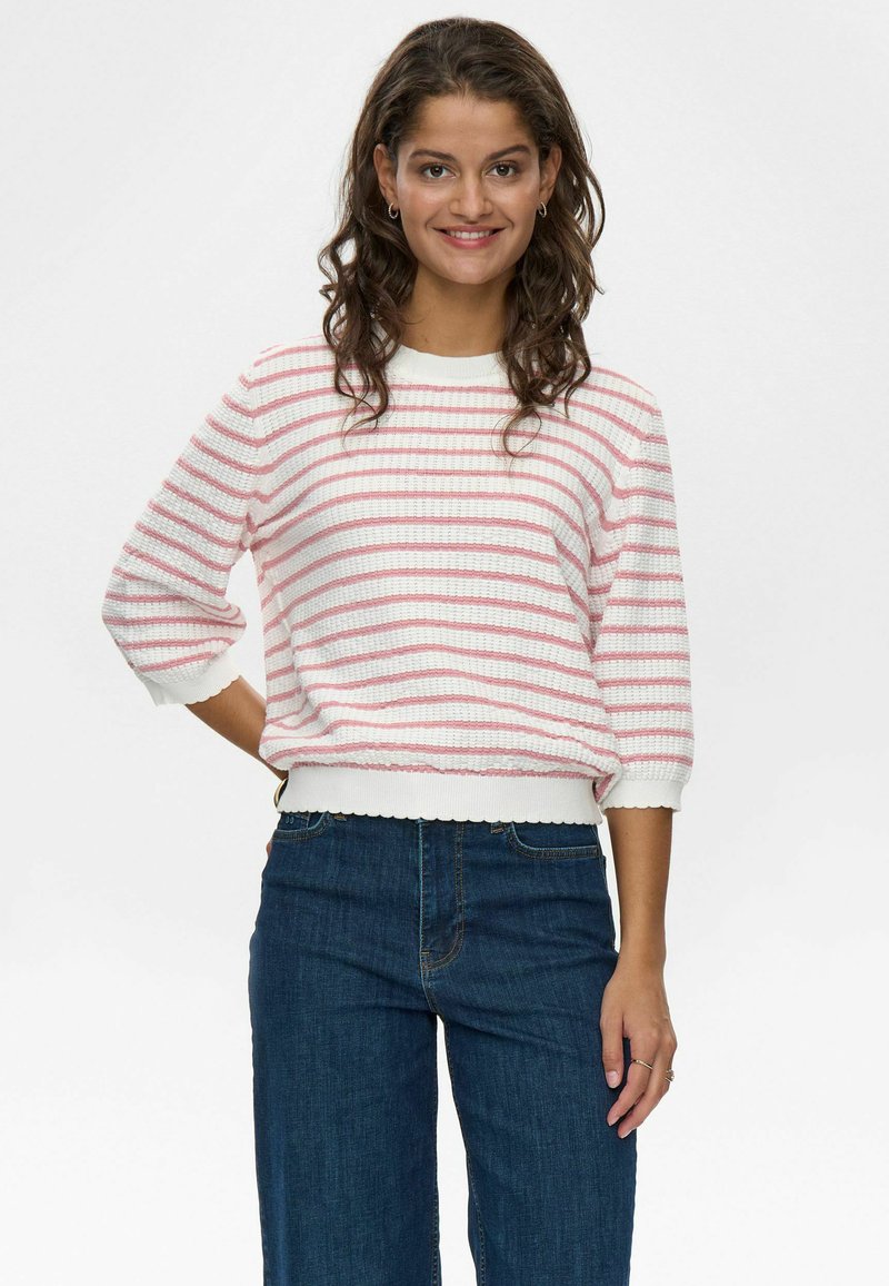 Csíkos pulóver fehér alapon rózsaszín csíkokkal, kötött textúrával, kerek nyakkivágással és háromnegyedes ujjal. Sötét kékdenim széles szárú nadrággal párosítva.