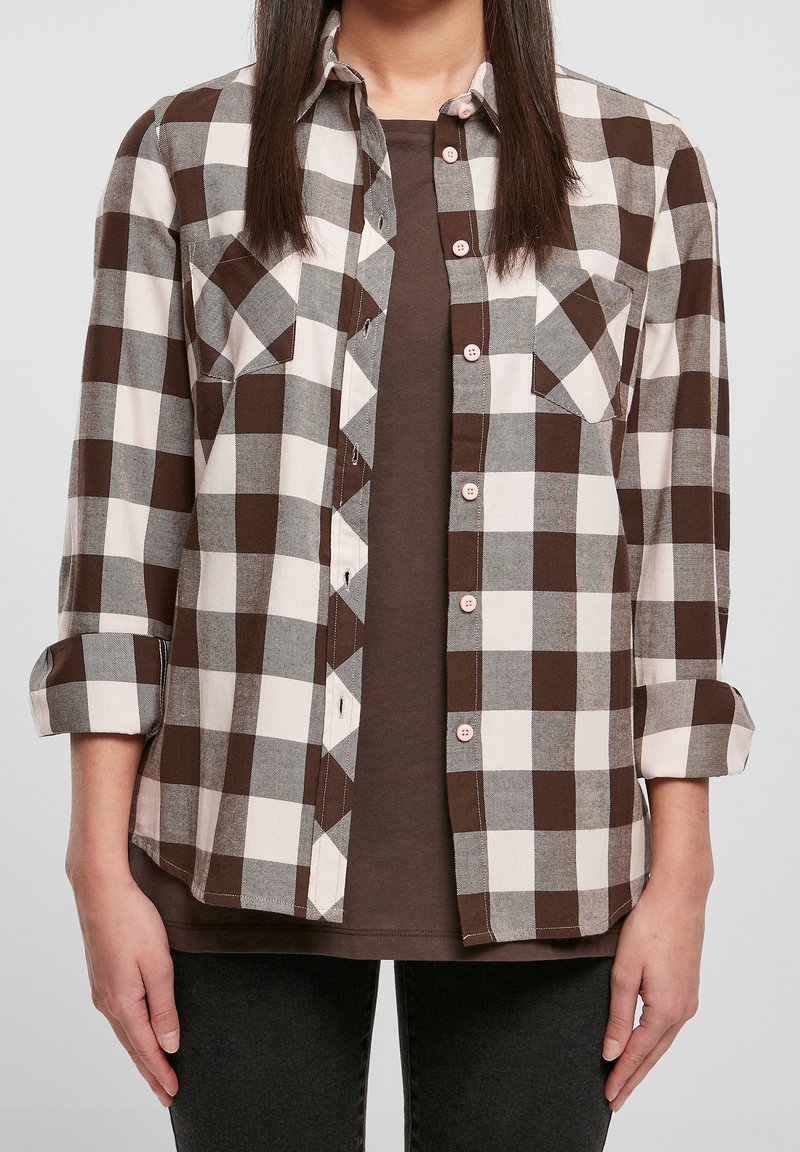 Femme portant une chemise à carreaux marron, blanc et gris boutonnée par-dessus un top marron, manches retroussées et pantalon noir.