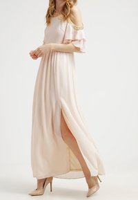 Vestido maxi de suave tela rosa claro con mangas de hombros al aire, ribetes en tono dorado y una abertura alta en el muslo. Combinado con tacones altos beige.