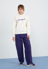 Kremeni dukserica s plavim "carhartt" logotipom, uparena s širokim tamnoljubičastim hlačama i ljubičastim tenisicama s bijelim prugama.