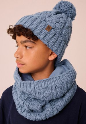 Stickad blå mössa med en pom-pom och matchande halsduk, med en strukturerad kabelmönster och en liten mörk etikettdetalj.