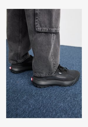 Czarne sportowe buty z teksturowanymi podeszwami, siateczkową cholewką i wyraźnymi czerwonymi akcentami, zestawione z luźnymi szarymi spodniami, na niebieskiej wykładzine.