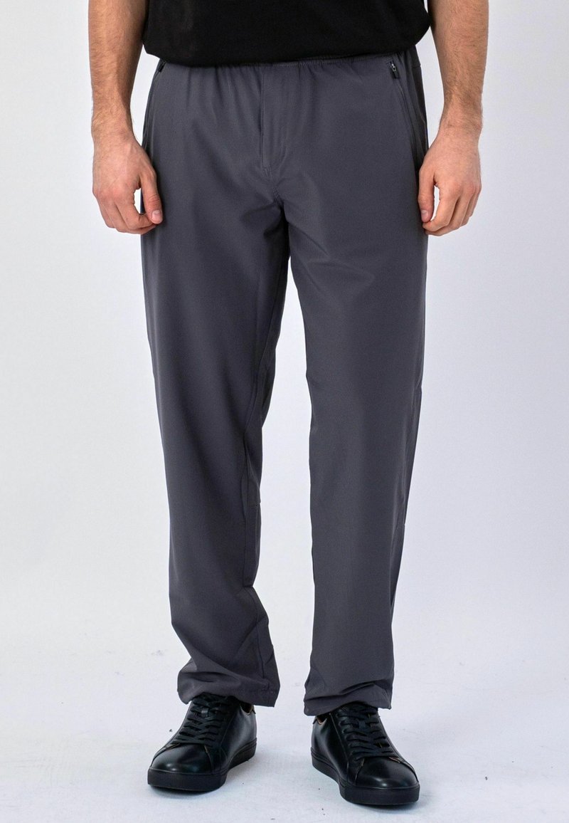 Homme portant un pantalon décontracté gris foncé et des baskets noires, debout devant un fond blanc.