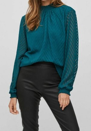 Femme portant un chemisier transparent bleu sarcelle à manches longues avec un motif chevrons texturé et un pantalon en cuir noir ajusté sur un fond neutre.