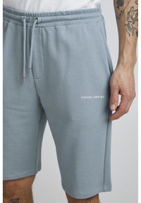 Shorts en coton léger bleu clair, avec une taille élastique, un cordon de serrage et le texte imprimé "CASUAL" sur la jambe gauche.