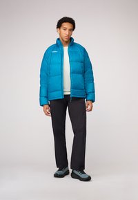 Doudoune teal avec un col haut, présentant des sections matelassées. Assortie à un pantalon noir et des chaussures de randonnée teal-noir. Fond uni.