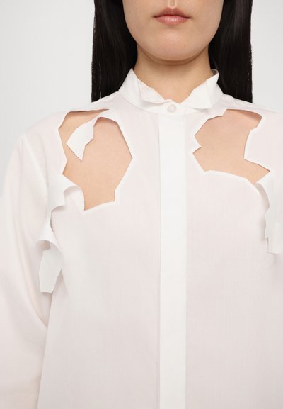 White cotton shirt featuring unique cut-out sections at the shoulders, ruffled collar, and a buttoned front placket.