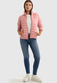 Heledus roosa puffjakker horisontaalse õmblusega, kõrge kraega ja küljetaskutega, kantud valge särgi peal ja siniste teksadega.