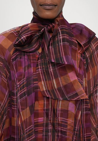 Geruite blouse van doorzichtige, lichtgewicht stof in paarse, bruine en oranje tinten. Heeft een grote strik bij de halslijn.