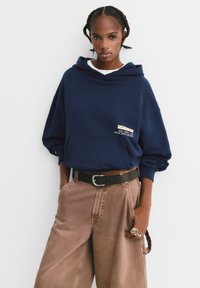 Námornícky oversized mikina s prednou vreckom, s nášivkou loga. V kombinácii so svetlohnedými nohavicami širokého strihu a čiernym opaskom.